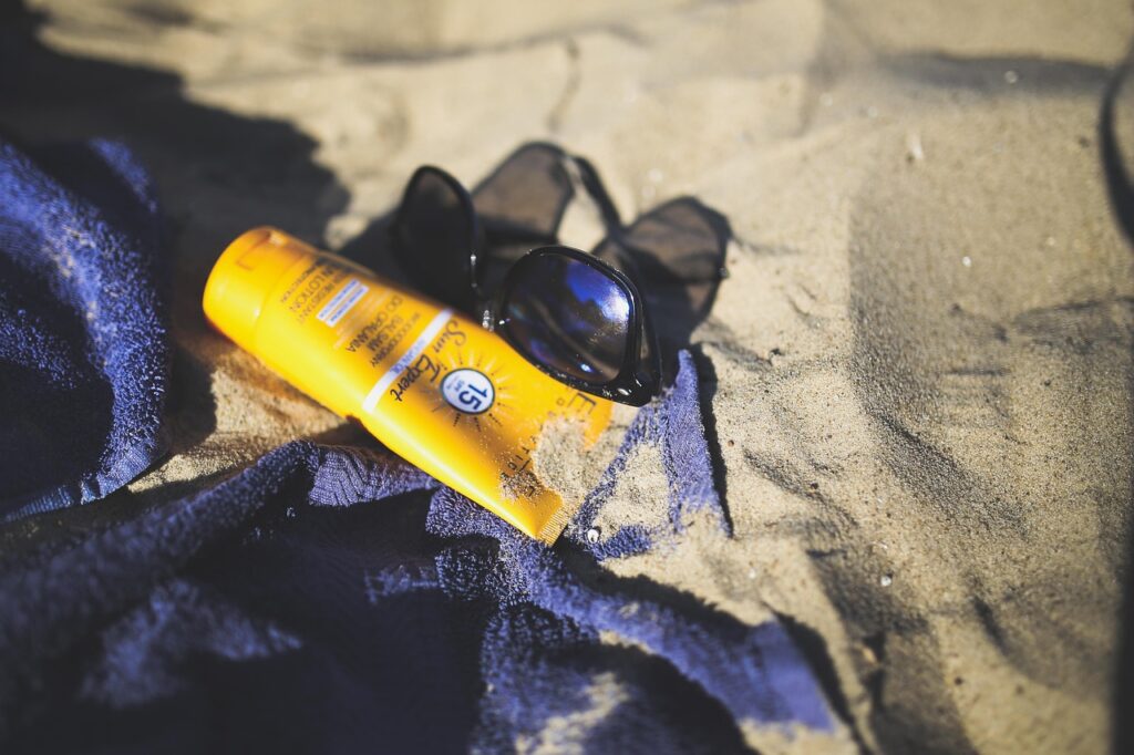Yellow sunscreen tube resting on a towel in sand beside sunglasses, representing avobenzone in sunscreens
