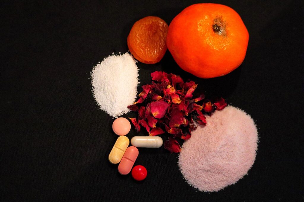 White and pink supplement powders beside capsules, dried fruit and an orange on a dark surface, representing collagen supplements