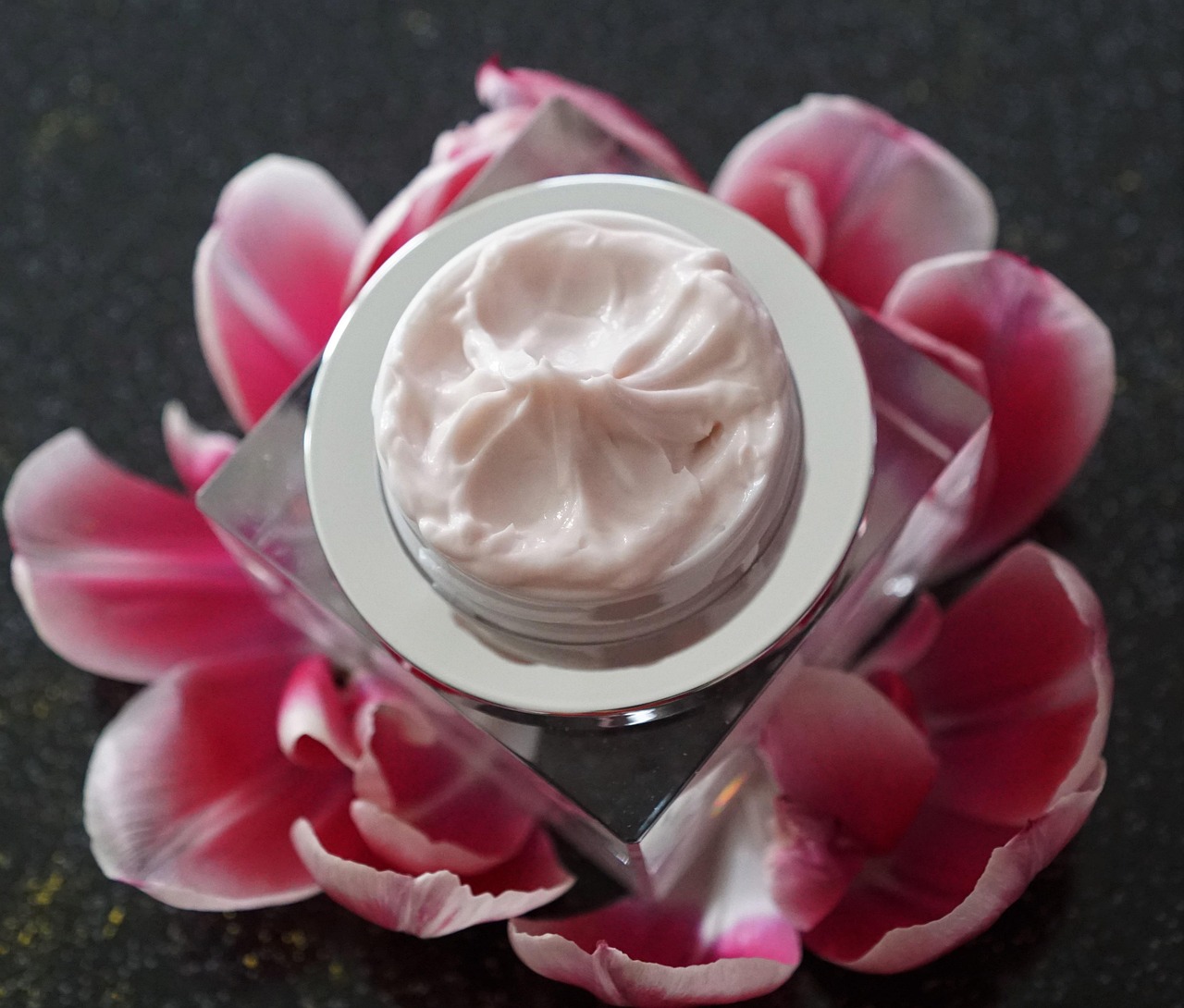 Jar of cream on a square glass base surrounded by pink petals, representing Differin gel topical retinoid treatment