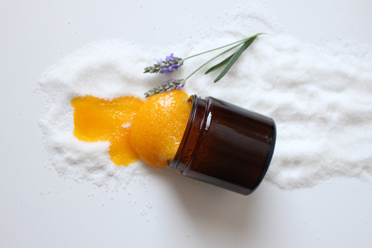 Amber jar tipped over spilling orange scrub onto white salt beside a lavender sprig, representing an exfoliating skincare product