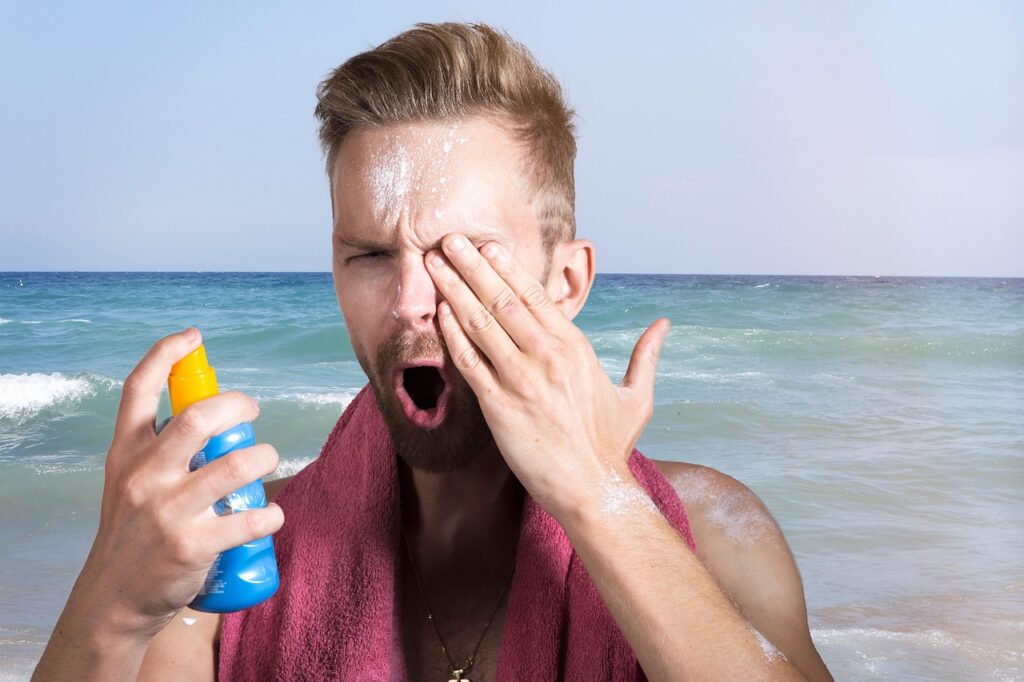 Man on a beach rubbing sunscreen on his face while holding a spray bottle, representing self-tanners and sun products