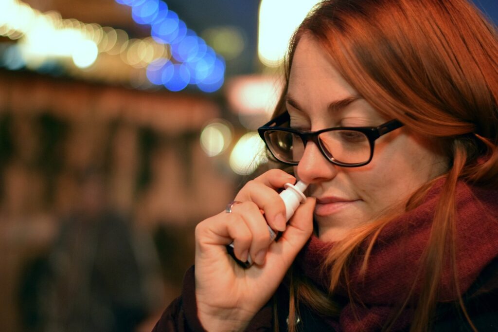 Woman in glasses and scarf using a nasal spray, representing decongestants like Sudafed