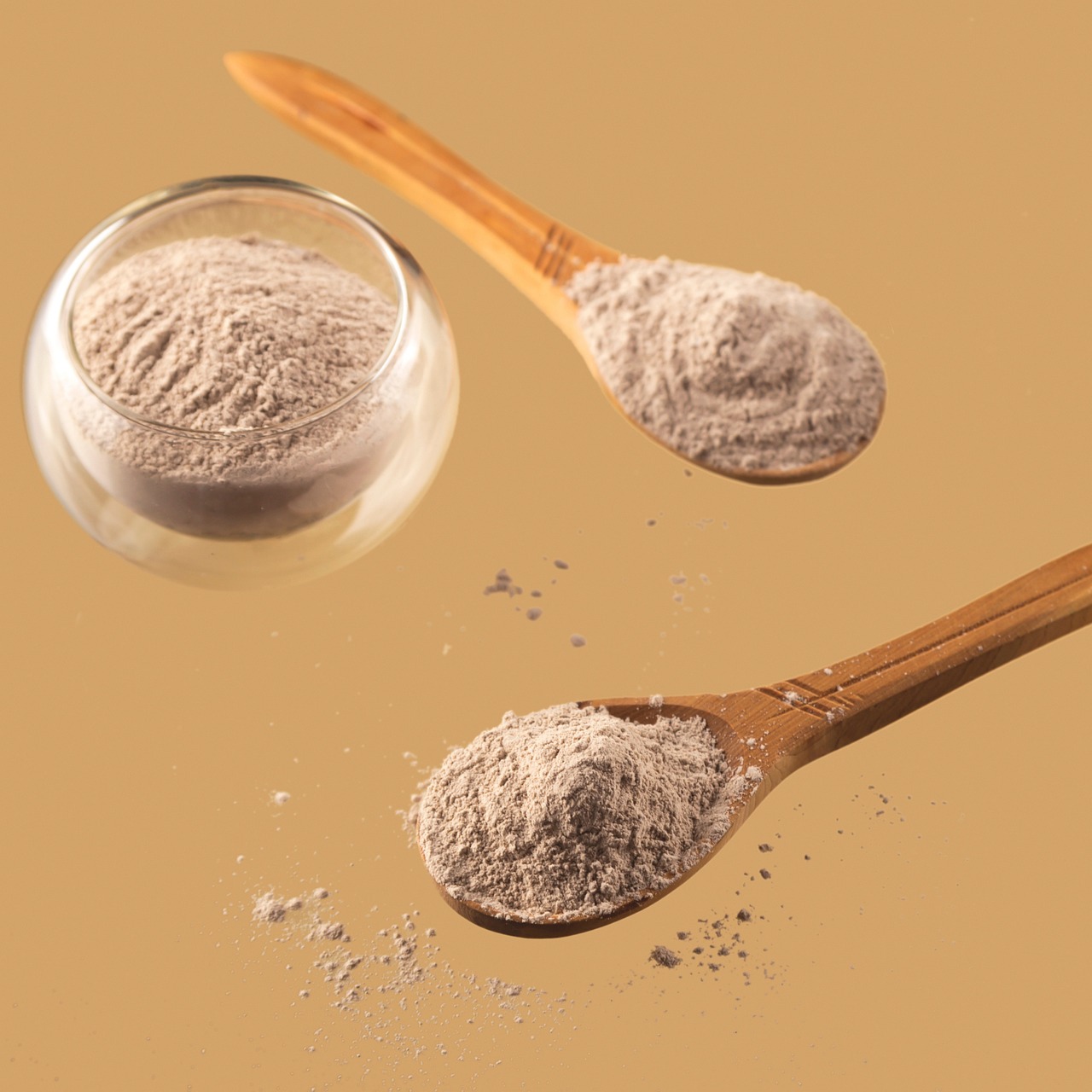 Pale mineral powder in a glass bowl and on a wooden spoon on a tan background, representing kojic acid skincare