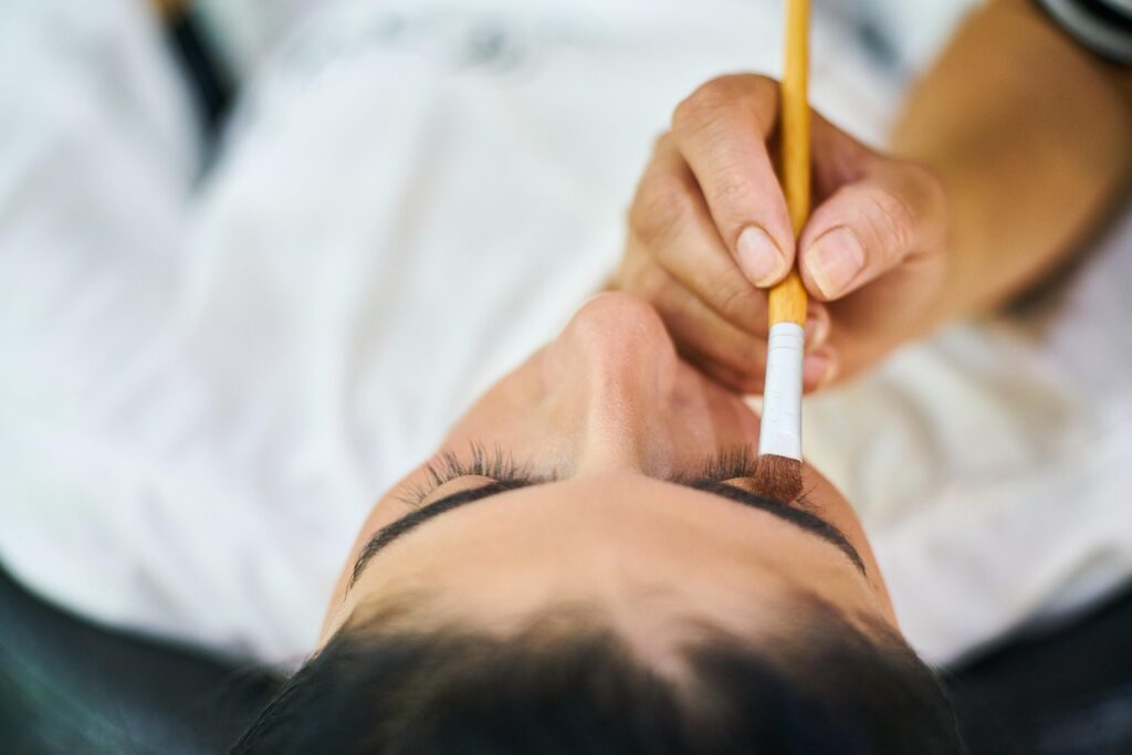 Beauty technician drawing on a reclining woman's eyebrows with a brush, illustrating brow cosmetic procedures like microblading
