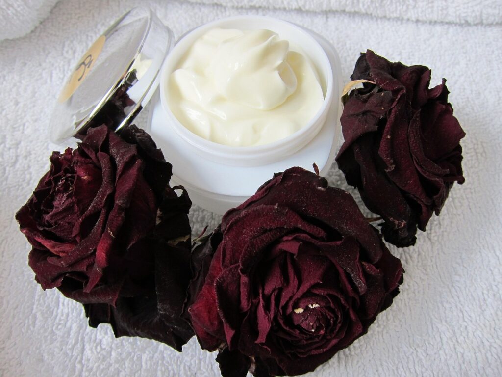 Jar of white cream beside dark red roses on a white towel, representing skin care and hair removal cream