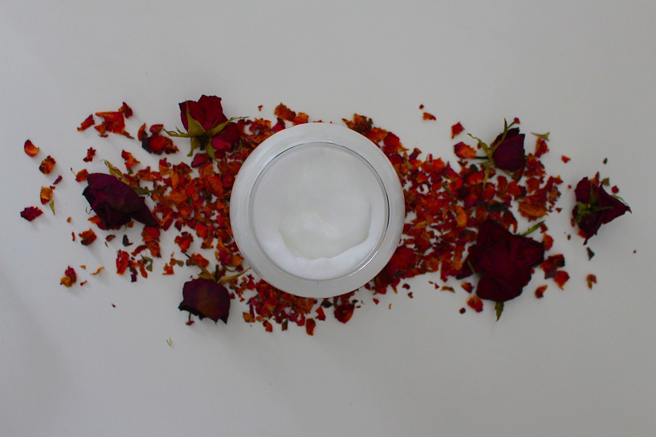 Round jar of white face cream on a pale surface scattered with dried rose petals, representing personal care products