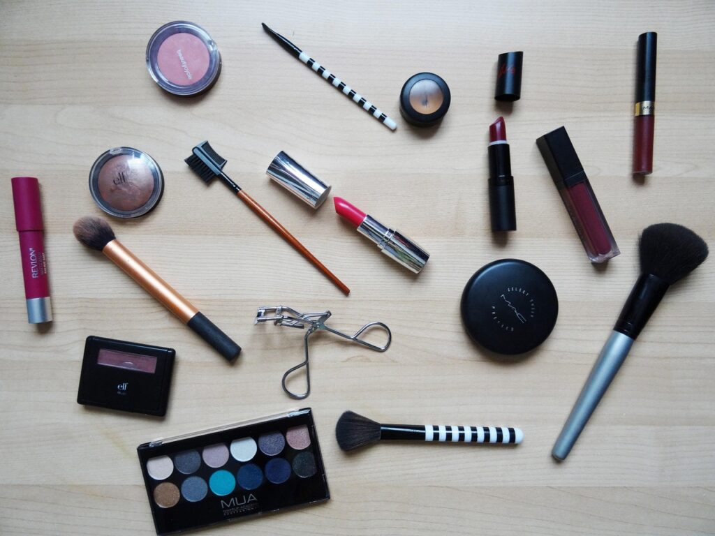 Flat lay of lipsticks, eyeshadow palettes, brushes and mascara on a light wooden table, representing pregnancy-safe makeup