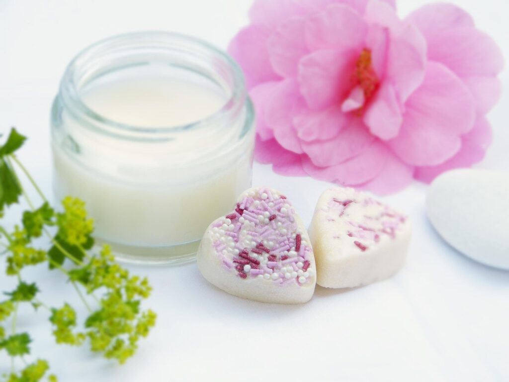 Glass jar of creamy shea butter beside a pink flower and small soaps on a white table