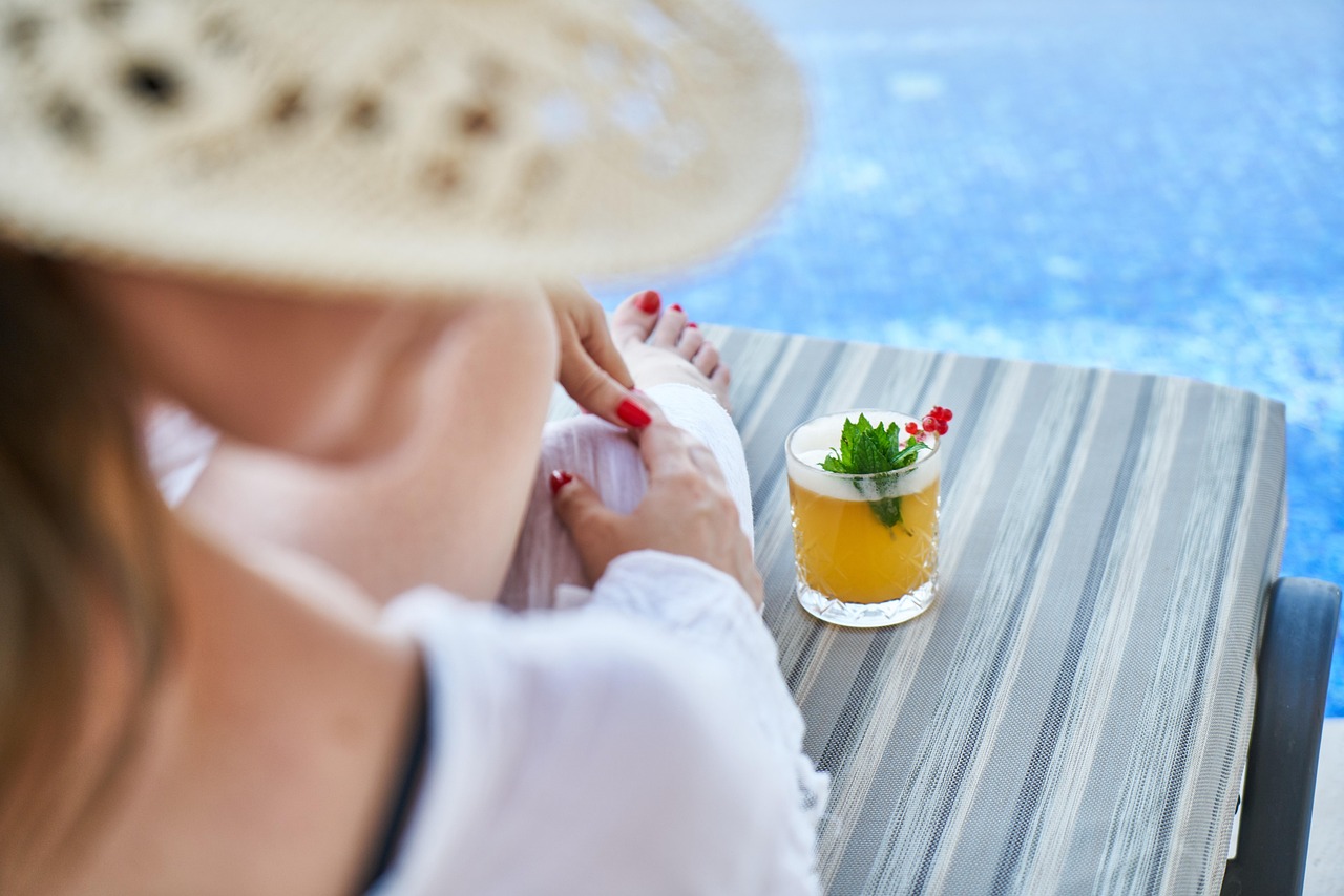 Woman in a sun hat relaxing at a poolside table by the ocean, representing summer pregnancy skincare