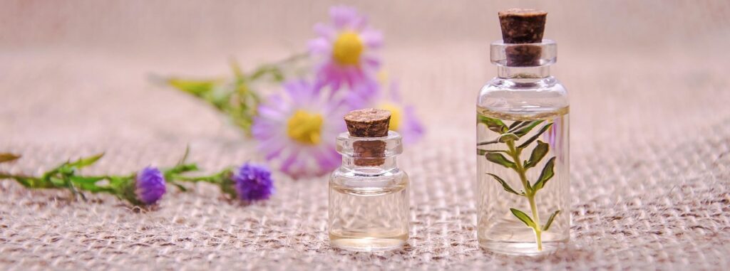 Two small corked glass bottles of herbal essential oil beside purple flowers on burlap