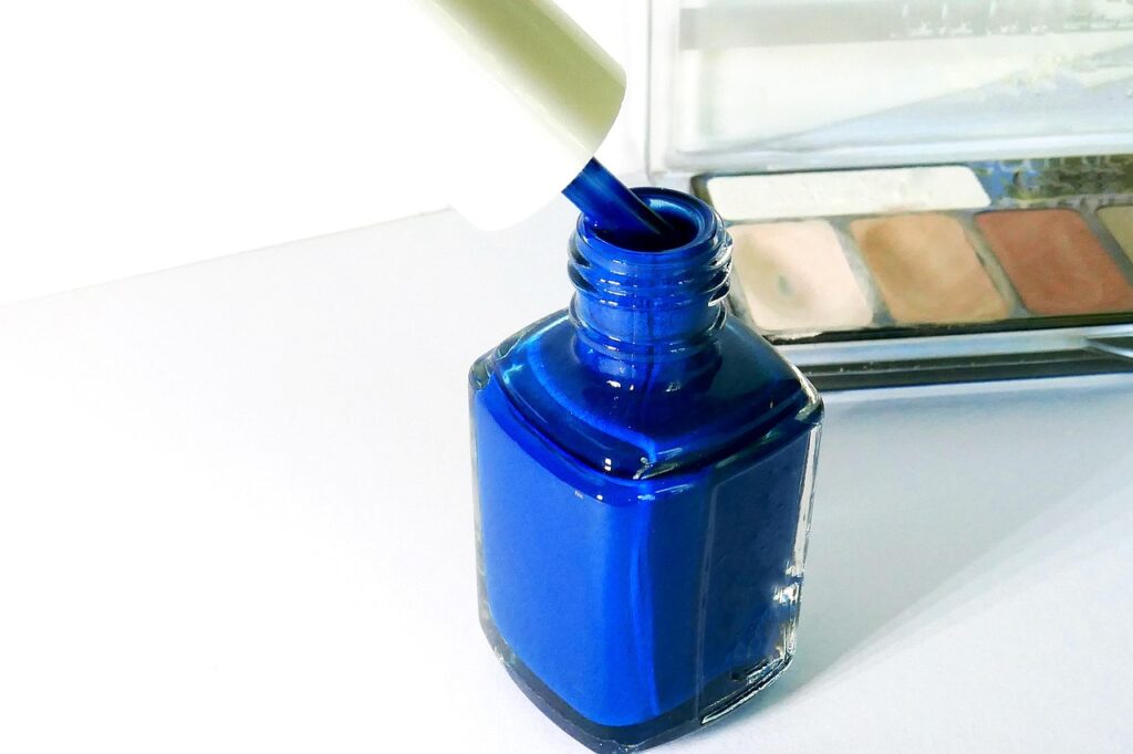 Open bottle of blue nail polish with a brush beside an eyeshadow palette, representing makeup and nail products