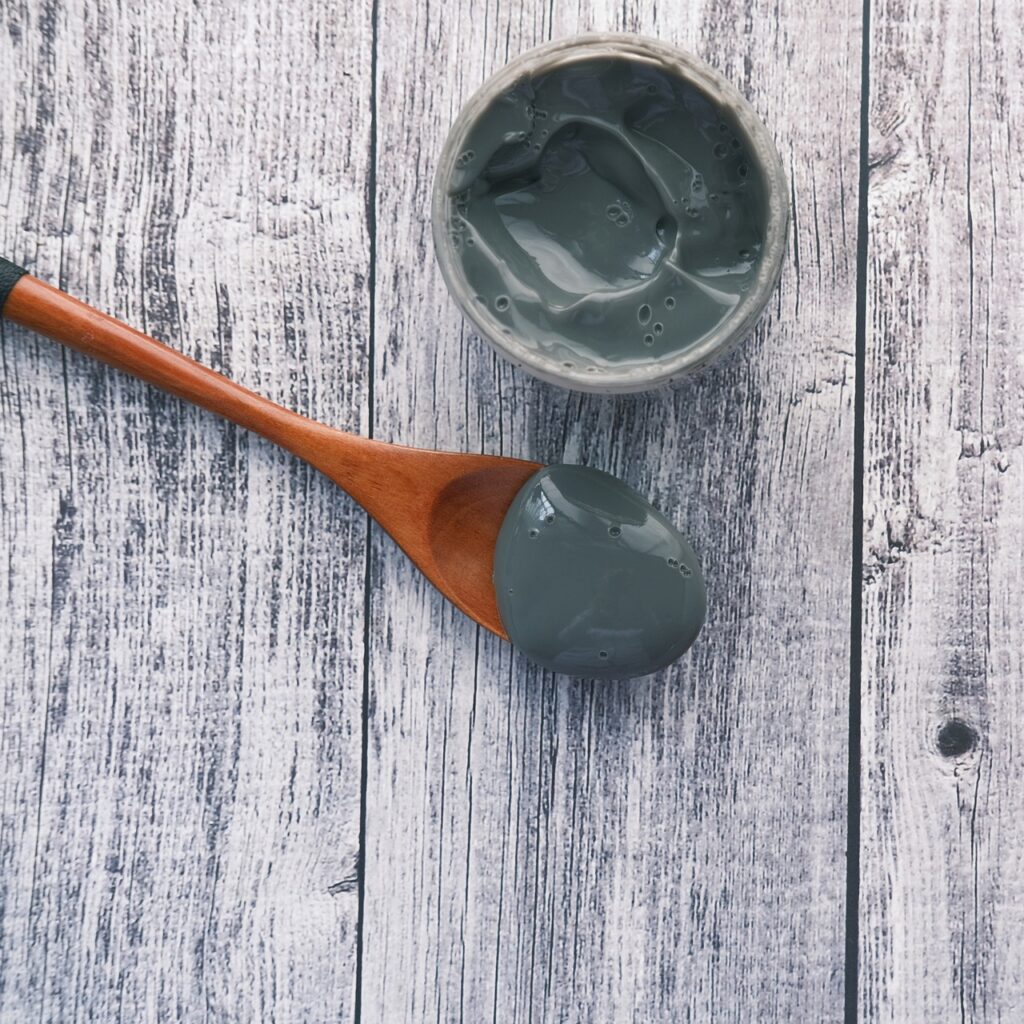 Bowl of gray clay face mask with a wooden spoon on a weathered wood surface, representing skincare