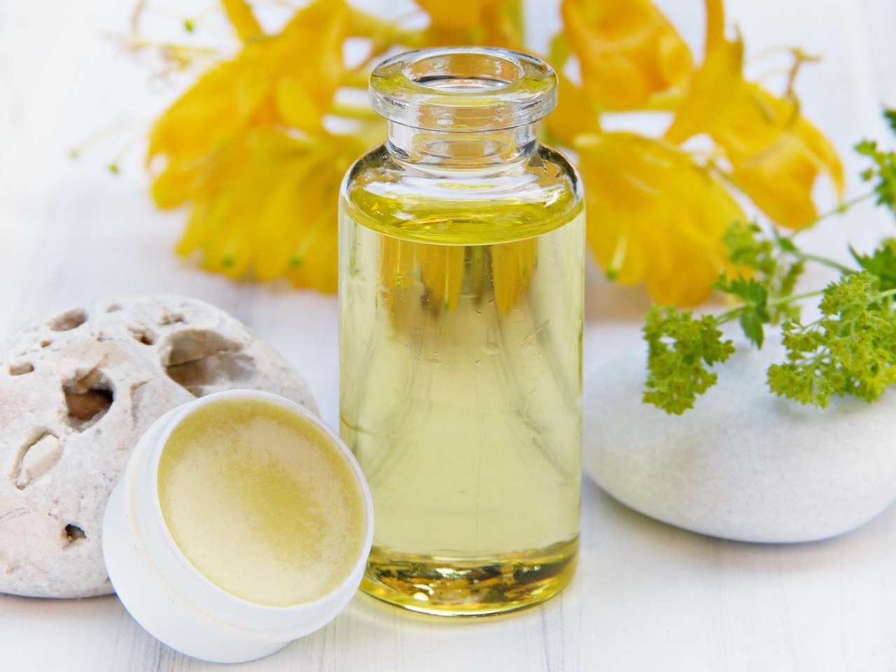 Clear bottle of oil beside a small tin of lip balm and yellow blossoms, representing gentle winter skincare