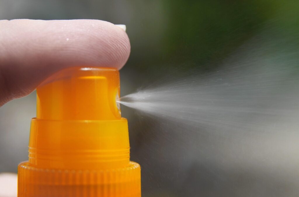 Finger pressing the pump of an orange sunscreen spray bottle releasing a fine mist, representing zinc oxide sun protection